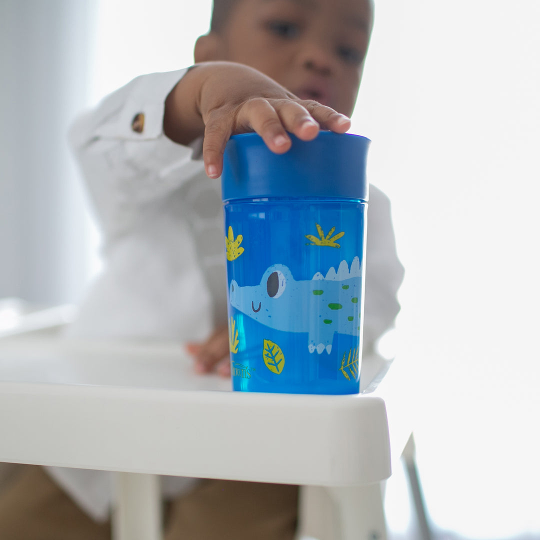 A toddler in a white shirt eagerly reaches for a blue Dr. Brown's® Milestones™ Cheers360™ Cup, which is decorated with a cartoon crocodile and leaves, resting on the high chair tray. The background is softly blurred.