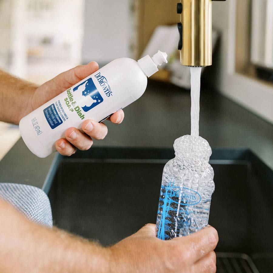 While standing at the kitchen sink, a person fills a reusable bottle with water from a brass faucet, holding Dr. Brown’s™ Bottle & Dish Soap—celebrated for its natural plant-derived ingredients—in the other hand. This scene highlights sustainability and eco-friendly choices.