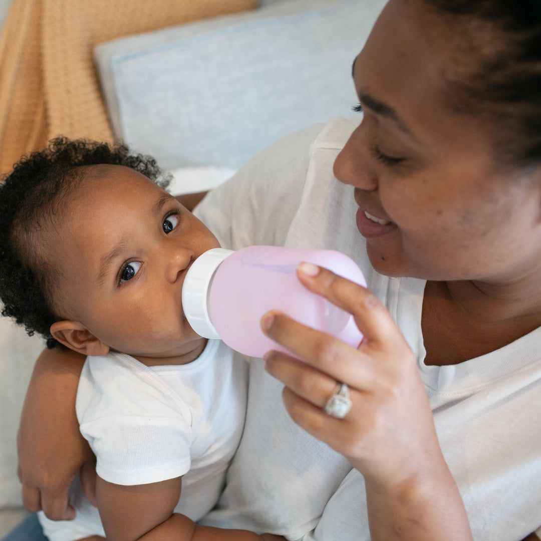 A woman with a joyful expression feeds a baby using a Dr. Brown's Natural Flow® Options+™ Wide-Neck Glass Bottle, adorned with a silicone sleeve. Both are clad in white shirts, comfortably seated amid a cozy ambiance with a soft blanket in the backdrop.