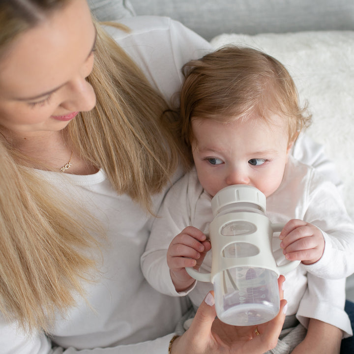 A woman with long blonde hair, wearing a white shirt, holds a baby who is skillfully drinking from the Dr. Brown’s® Milestones™ Wide-Neck Sippy Spout Bottle with Silicone Handles, 9 oz/270 mL. The baby, dressed in a white shirt and featuring light brown hair, looks content while being held on a soft, light-colored surface.