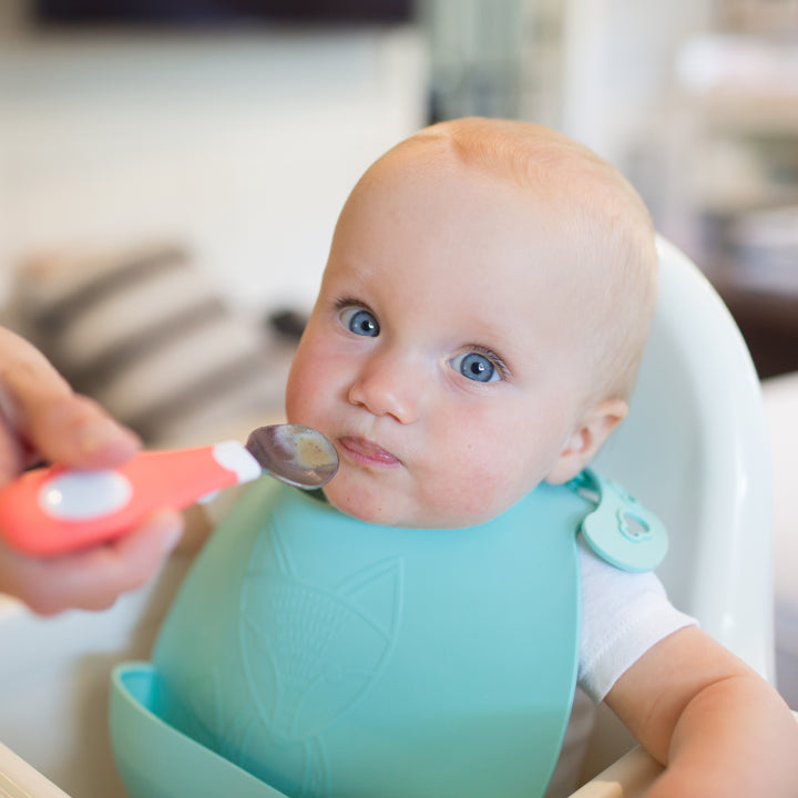 A baby with blue eyes sits in a high chair, wearing a teal bib. An adult hand holds Dr. Brown's Designed to Nourish Soft-Grip Spoon, which is nearing the baby's mouth. The background is softly blurred, showing a striped pillow.