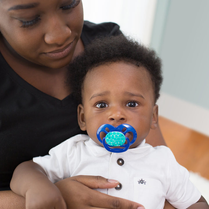 A baby, wearing a white shirt and using a Dr. Brown’s™ Advantage™ Pacifier from Dr. Brown's, sits on an adult's lap. The baby looks directly at the camera with the pacifier securely in their mouth, while the adult gazes down at them. The background is softly blurred.