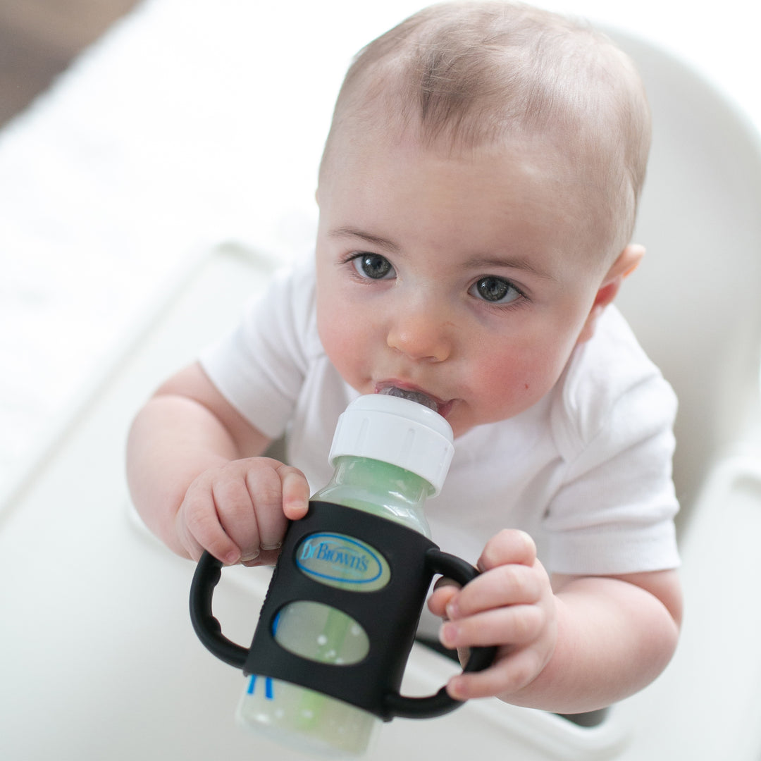 A baby in a white onesie sits in a high chair, holding Dr. Brown’s® Milestones™ Narrow Silicone Handles attached to a baby bottle. With a curious expression, the infant demonstrates impressive independent drinking skills.