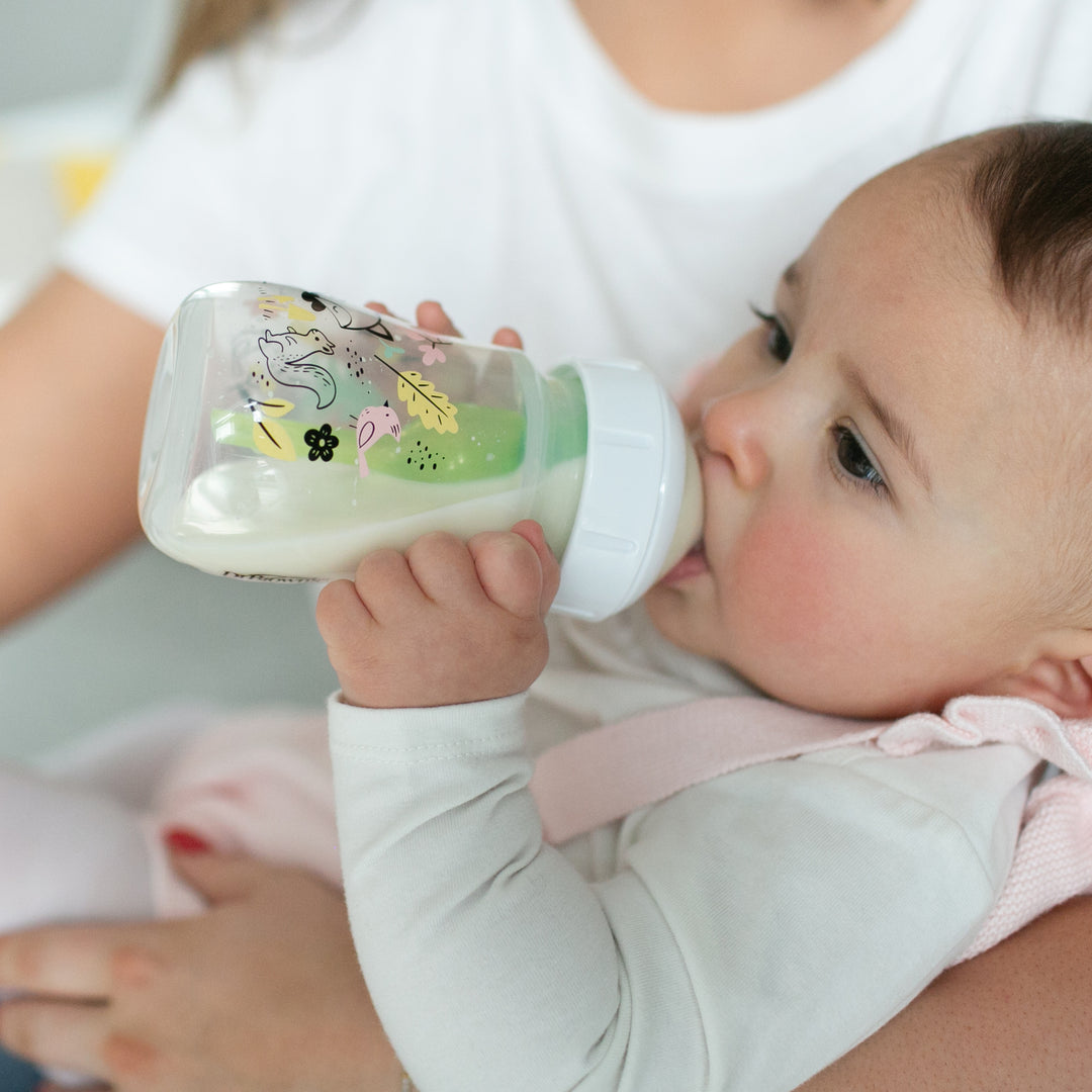 A baby in a white outfit is gently cradled in someone's arms, drinking from Dr. Brown's Natural Flow® Anti-Colic Options+™ Wide-Neck Baby Bottle, which features playful illustrations. The person holding the baby wears a white shirt and ensures comfort with the bottle’s slow flow nipple that mimics a breast-like feel.