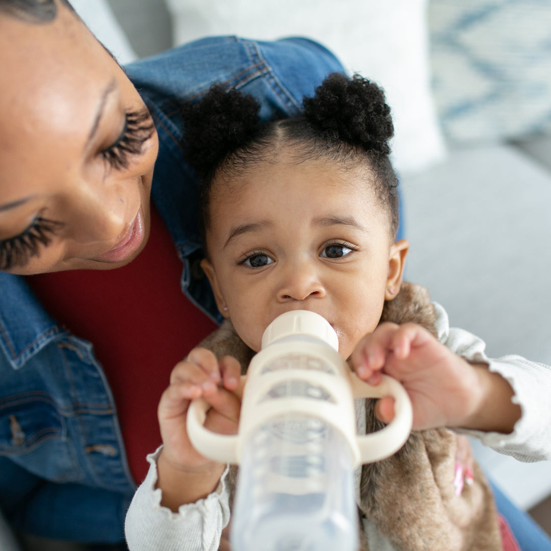 A woman in a denim jacket cradles a baby drinking from a Dr. Brown's® Milestones™ Narrow Sippy Spout Bottle with Silicone Handles. The little one, with two small puff ponytails, wears a light-colored top as they enjoy the serene moment together in a cozy indoor setting.