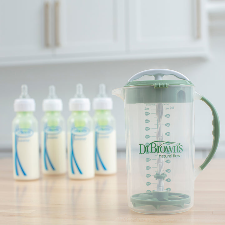 A Dr. Brown's Natural Flow® Formula Mixing Pitcher with a green handle rests on a wooden surface. In the background, four baby bottles filled with milk are aligned and slightly out of focus. The scene is set in a kitchen with light-colored cabinets, ideal for effortless formula preparation.