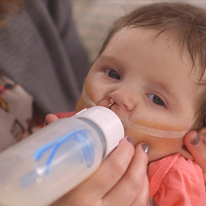 A baby with nasal tubes is being gently fed milk using a Dr. Brown’s™ Medical – Narrow Bottle Nipples. The little one, dressed in a pink outfit, nestles comfortably in the super soft embrace of an adult.