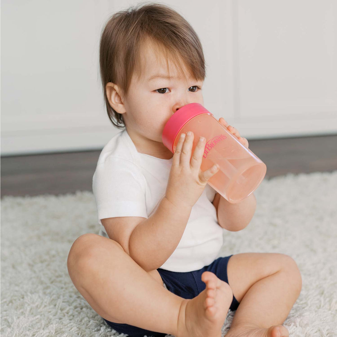 A toddler in a white shirt and blue shorts practices sipping skills with Dr. Brown's® Milestones™ Straw to 360° Cup Set.