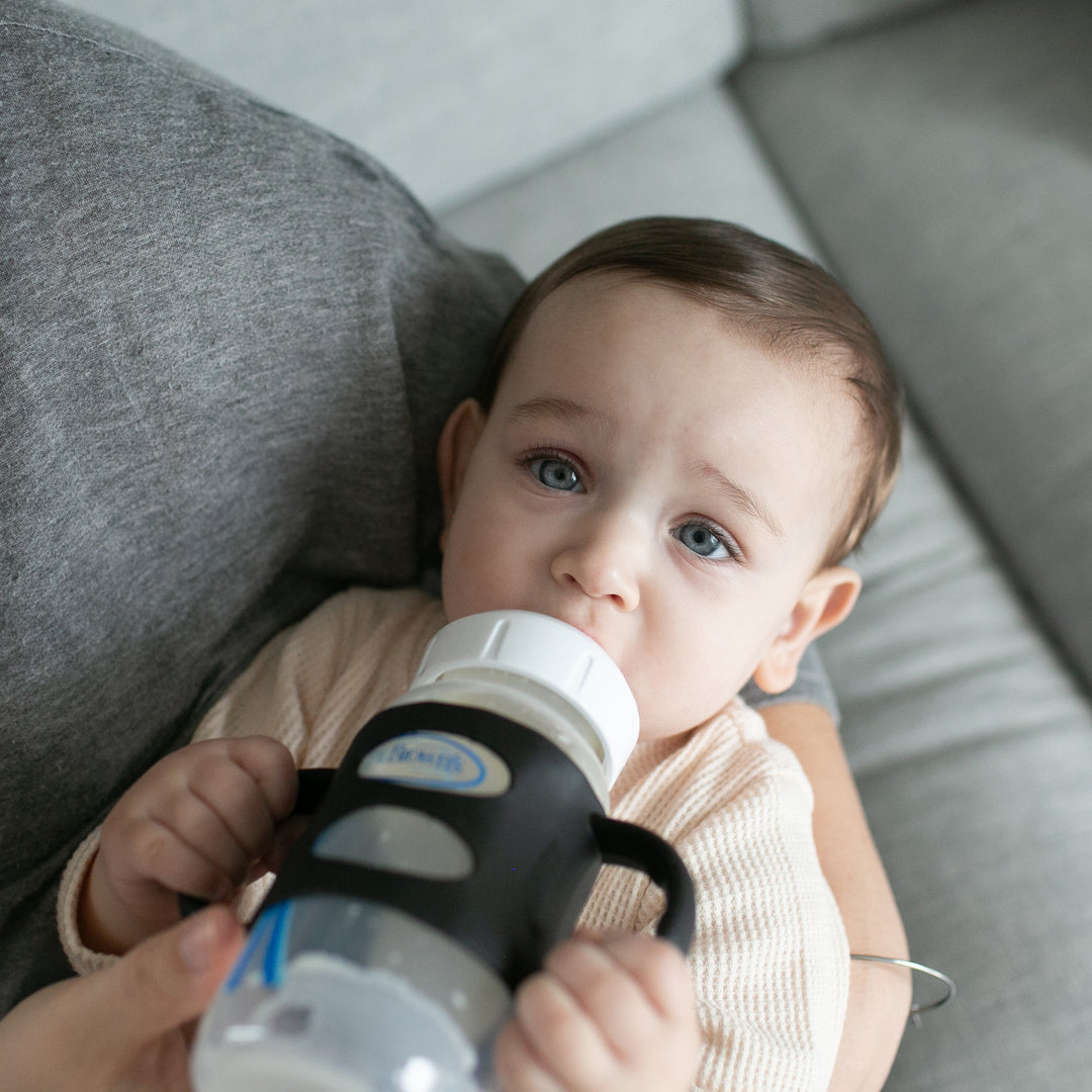 A baby with dark hair lies on a gray sofa, firmly holding a bottle with both hands using the Dr. Brown’s® Milestones™ Wide-Neck Silicone Handles. Dressed in a light-colored outfit, the baby looks up confidently while enjoying independent drinking.