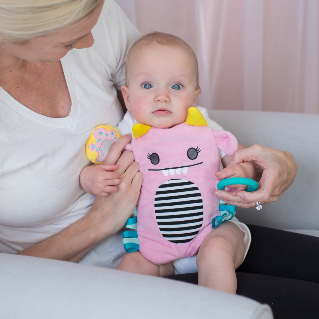 In a cozy room, a baby sits on an adult's lap, holding a toy and wearing an adorable pink monster-themed bib featuring yellow horns and black-and-white stripes. The adult, dressed in a white shirt, gently holds the baby's hands while using Dr. Brown’s™ Gripebelt™, a soothing heated tummy wrap from Dr. Brown's, to offer relief from colic.