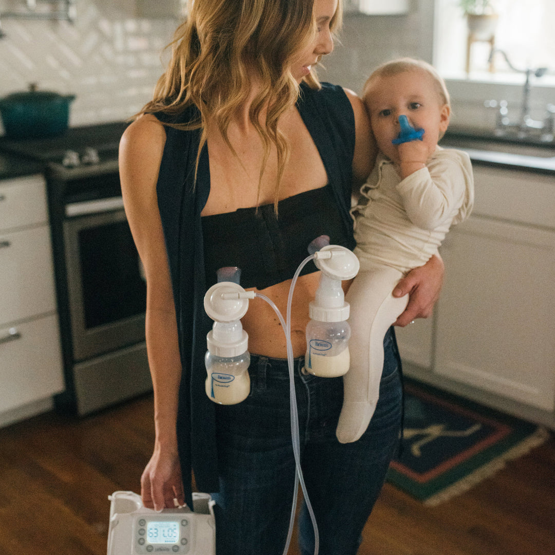 A woman stands in a kitchen holding a baby, effortlessly using an electric pump with two bottles partially filled with milk. Thanks to her Dr. Brown's Hands Free Pumping Bra from Dr. Brown's, she enjoys a custom fit while the baby, dressed in white, plays with a blue toy.