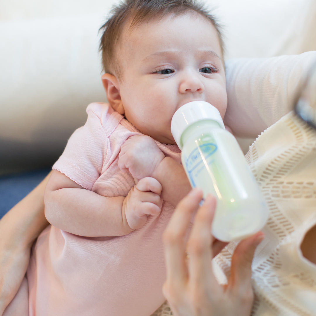 A baby wearing a pink onesie is being fed with Dr. Brown’s Natural Flow® Anti-Colic Options+™ Narrow Baby Bottle, which features a Level 1 Slow Flow Nipple designed to mimic breastfeeding, cradled gently in the arms of an adult. The cozy, intimate setting conveys the nurturing comfort and natural flow associated with this product by Dr. Brown's.