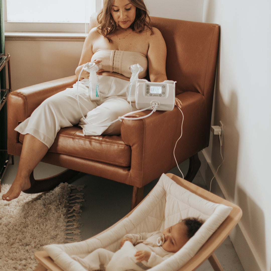 A woman sits in a brown armchair using an electric pump, glancing at a baby sleeping in a beige rocker nearby. The room feels serene with its soft, warm tone and woven rug. Her Dr. Brown's Hands Free Pumping Bra provides comfort as she tends to her little one with ease.