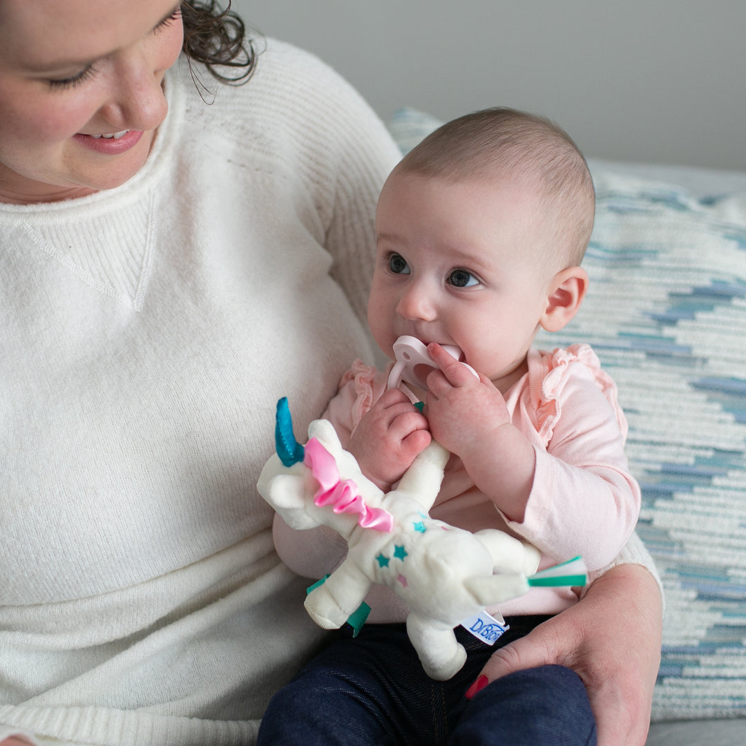 A woman in a white sweater beams as she holds a baby on her lap. The baby, dressed in a pink outfit, grips Dr. Brown’s® Lovey Pacifier and Teether Holder, Unicorn, along with a stuffed unicorn. They sit on a patterned cushion, sharing a moment of pure joy and comfort.