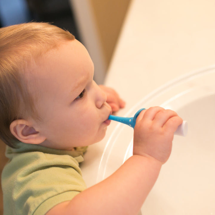 A toddler wearing a green shirt diligently brushes their teeth with Dr. Brown's™ Infant-to-Toddler Toothbrush, Elephant, 1-Pack, staying focused on maintaining oral hygiene at the bathroom sink against the softly lit background.