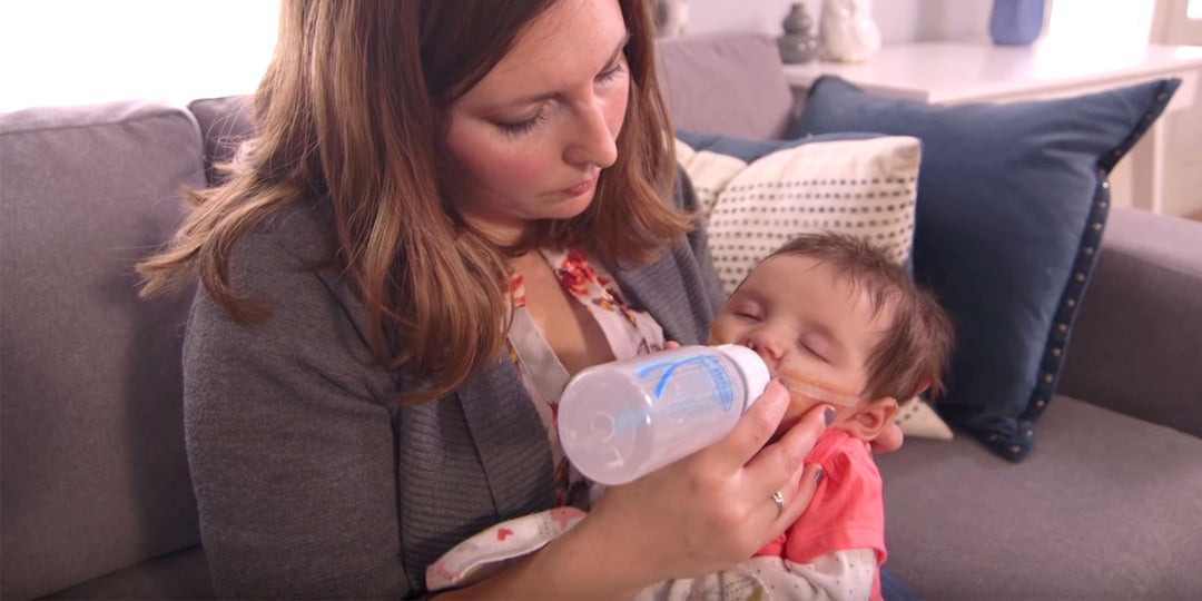 A woman with brown hair is sitting on a couch, gently feeding a baby with a bottle. The baby is wearing a pink outfit and has a feeding tube attached. Blue and patterned pillows are in the background.