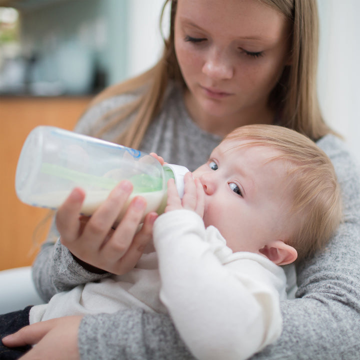 A woman with long hair cradles a baby in her lap, gently feeding them using Dr. Brown’s Natural Flow® Options+™ Wide-Neck Bottle from the Replacement Vent Kit. The baby, dressed in a white outfit, gazes upwards while drinking. A softly blurred background ensures the tender moment remains the center of attention.