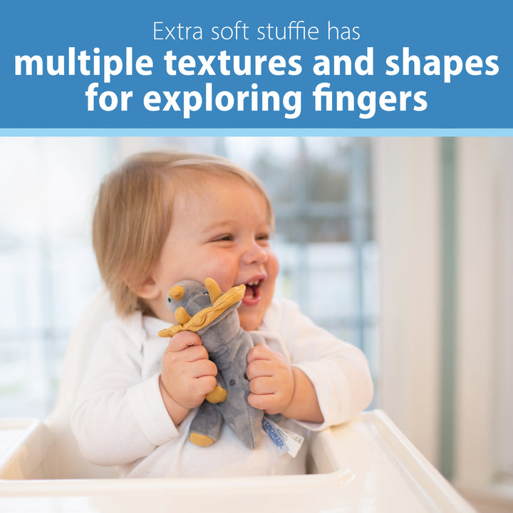 A beaming baby sits in a high chair, holding Dr. Brown’s® Lovey Pacifier and Teether Holder, Triceratops—a plush toy with multiple textures and shapes for curious fingers to explore. The text above reads, "Extra soft stuffie has diverse textures and shapes for exploring fingers.