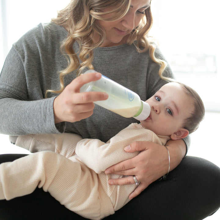 A woman with wavy hair is sitting and feeding a baby using the Dr. Brown’s Natural Flow® Anti-Colic Options+™ Narrow Glass Baby Bottle, which features a Level 1 Slow Flow Nipple. The baby, dressed in a light-colored onesie, is cradled in her arms. The softly blurred background suggests a cozy indoor environment, promoting an anti-colic experience.