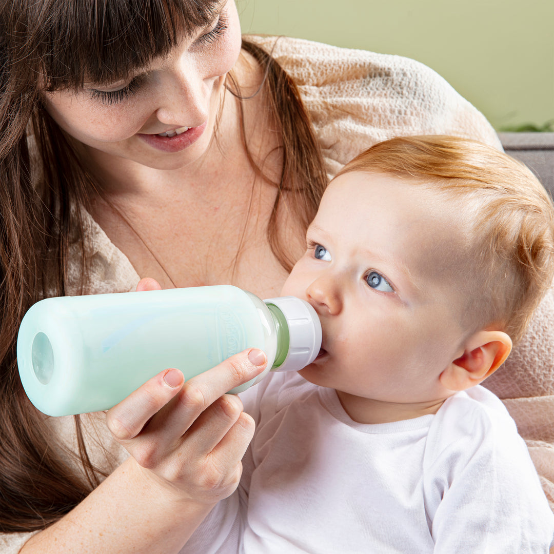 A woman with long brown hair smiles at a baby as she feeds him from a Dr. Brown’s Natural Flow® Options+™ Narrow Glass Bottle. The baby, dressed in a white shirt, looks upwards while drinking. This nurturing moment is warmly captured, enhanced by the protective silicone sleeve on the bottle from Dr. Brown's.