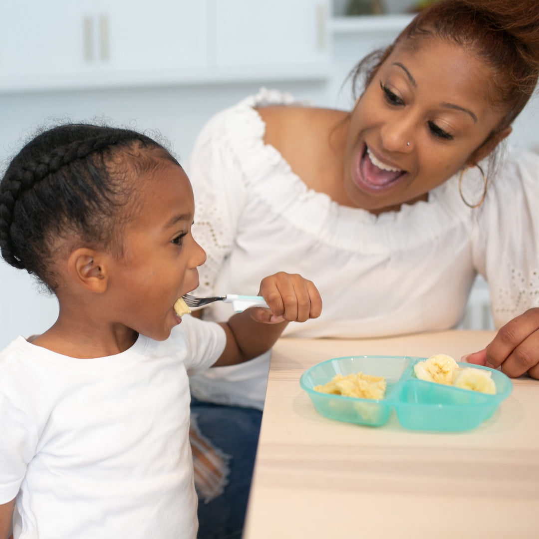 A woman and a young child sit at a table, smiling. The child enjoys their toddler meal from Dr. Brown's™ Divided Plate, which is light blue in color, while holding a spoon. The woman, dressed in a white top with her hair tied back, watches lovingly as the scene unfolds in the warm ambiance of a bright kitchen setting.