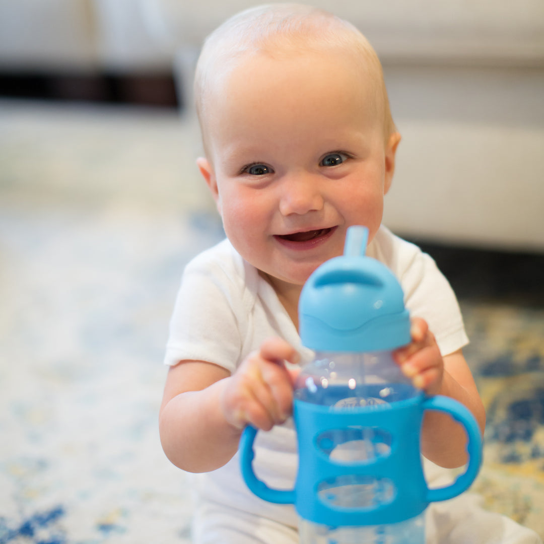 A smiling baby, dressed in a white onesie, holds Dr. Brown’s™ Milestones™ Wide-Neck Sippy Straw Bottle with Silicone Handles, showcasing their independent drinking skills. The scene features the baby seated on a plush, patterned carpet with a sofa softly blurred in the background.