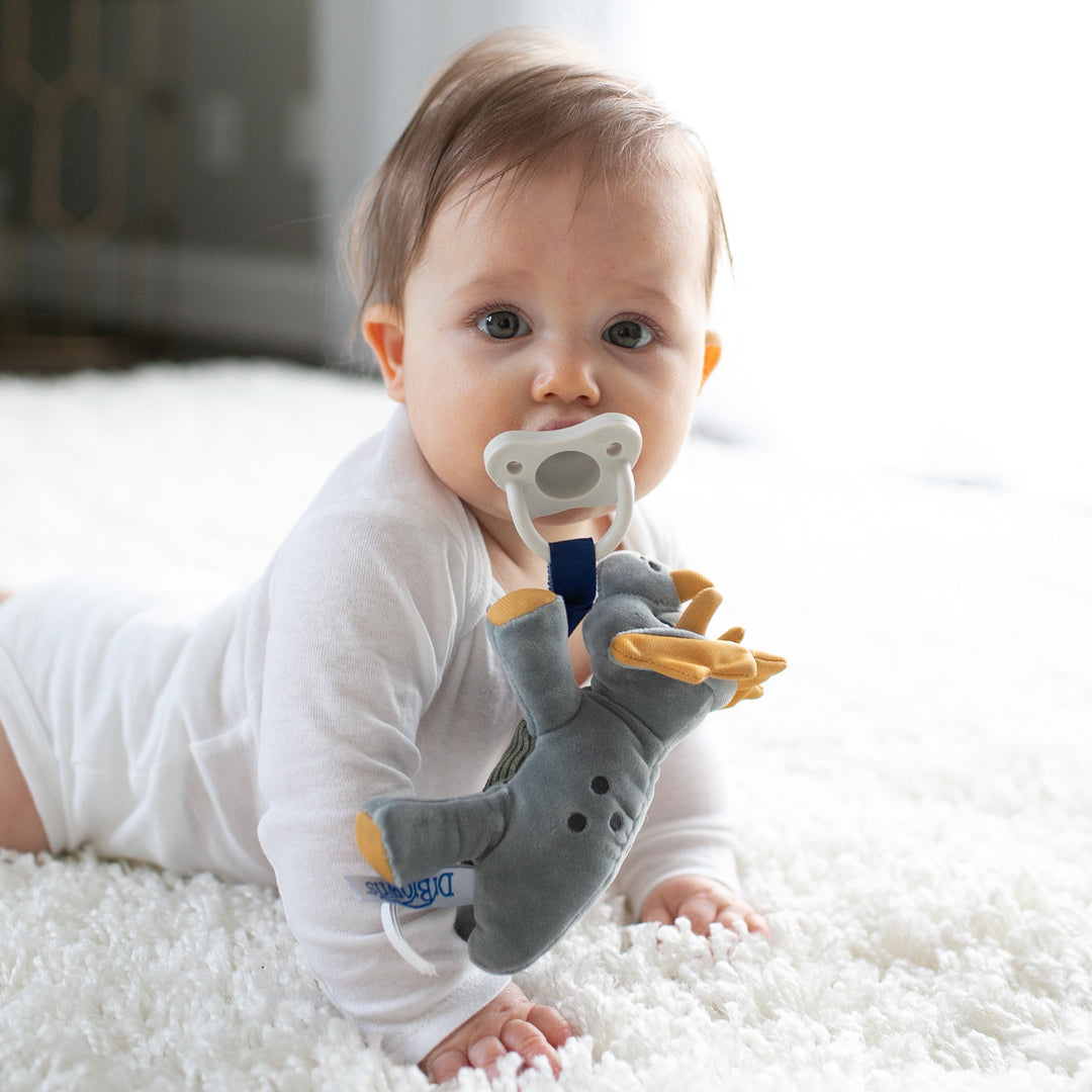 A baby, dressed in a white onesie, is lying on a fluffy white rug and sucking on the pacifier from Dr. Brown’s® Lovey Pacifier and Teether Holder. Attached to the pacifier is a plush triceratops, which doubles as both a teether holder and comforting lovey toy, playfully dangling within reach of the baby. The softly lit room provides a gentle backdrop.