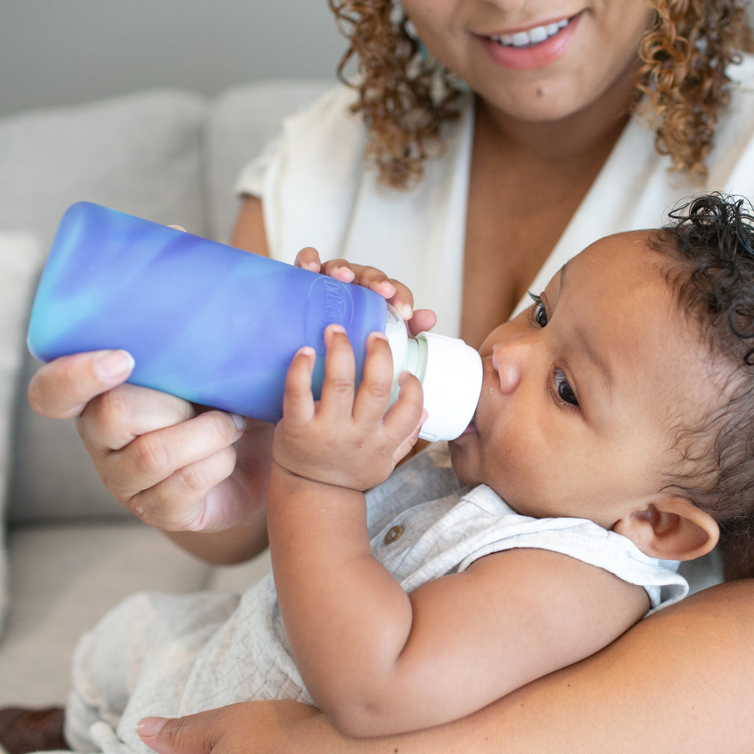 A caregiver with curly hair smiles gently while bottle-feeding a baby using Dr. Brown's Natural Flow® Options+™ Narrow Glass Bottle Silicone Sleeves. The baby lies comfortably on their lap, holding the blue bottle with both hands as they sit on a light-colored sofa.