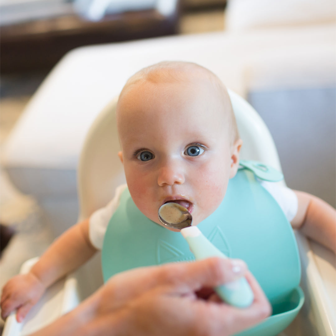 A baby in a high chair is being fed with a Dr. Brown's Designed to Nourish Soft-Grip Spoon from the toddler silverware set. The baby, wearing a light blue bib, has a focused expression as an adult hand approaches with the Dr. Brown’s stainless steel spoon.