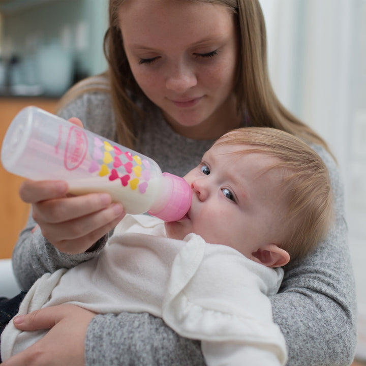 A young woman is sitting and bottle-feeding a baby with Dr. Brown's Natural Flow® Anti-Colic Options+™ Narrow Sippy Bottle Starter Kit, 8oz/250mL, in pink and white. The baby, wrapped in a white blanket, looks content. The softly blurred background highlights the tender moment shared between them.