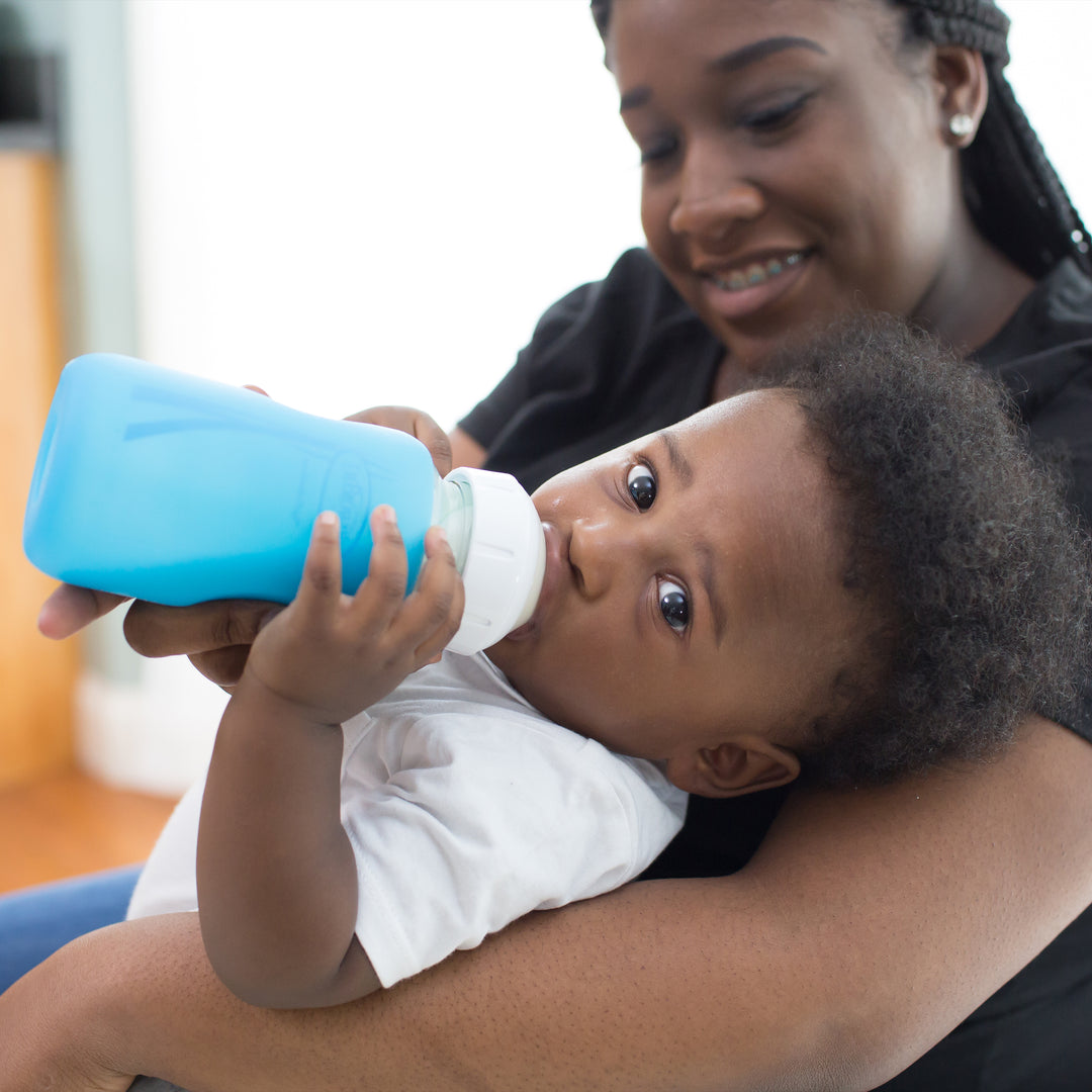 A baby effortlessly drinks from a blue Dr. Brown's Natural Flow® Options+™ Wide-Neck Glass Bottle Silicone Sleeve while being cradled by a smiling woman. Indoors, the soft lighting creates a warm and nurturing atmosphere, highlighting the baby's contentment and tranquility.