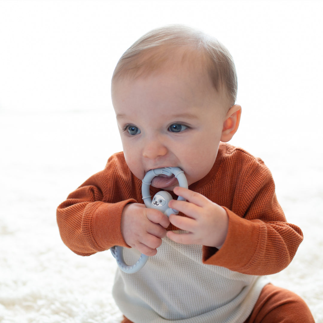 A baby with light brown hair is sitting on a soft, white carpet, wearing a rust-colored and cream outfit. The baby joyfully chews on Dr. Brown's™ Flexees™ Sloth Teether featuring a smiley face. The background is bright, suggesting natural light.