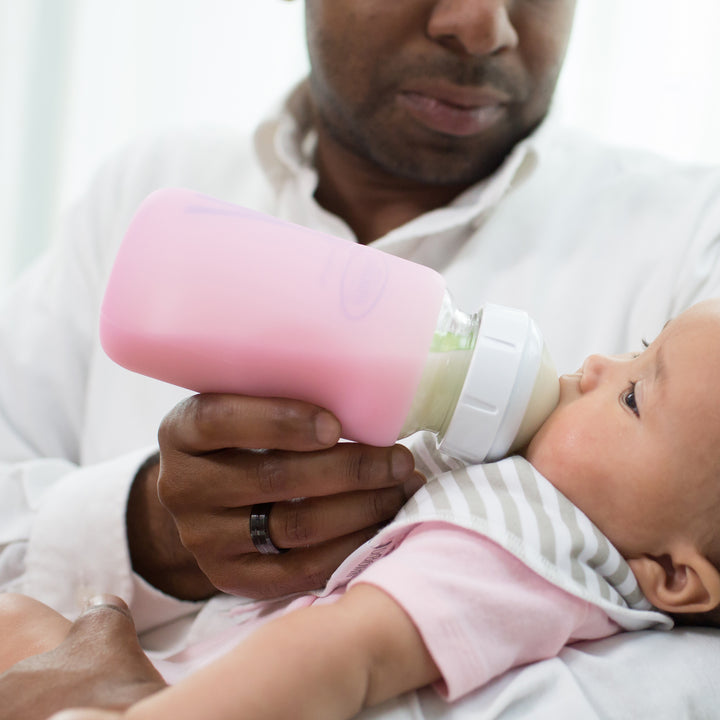 A man in a white shirt feeds an infant wearing a striped bib and pink outfit with a Dr. Brown's Natural Flow® Options+™ Wide-Neck Glass Bottle Silicone Sleeves. The bottle, featuring a white cap, stands out softly blurred in the background, beautifully capturing this intimate feeding moment.