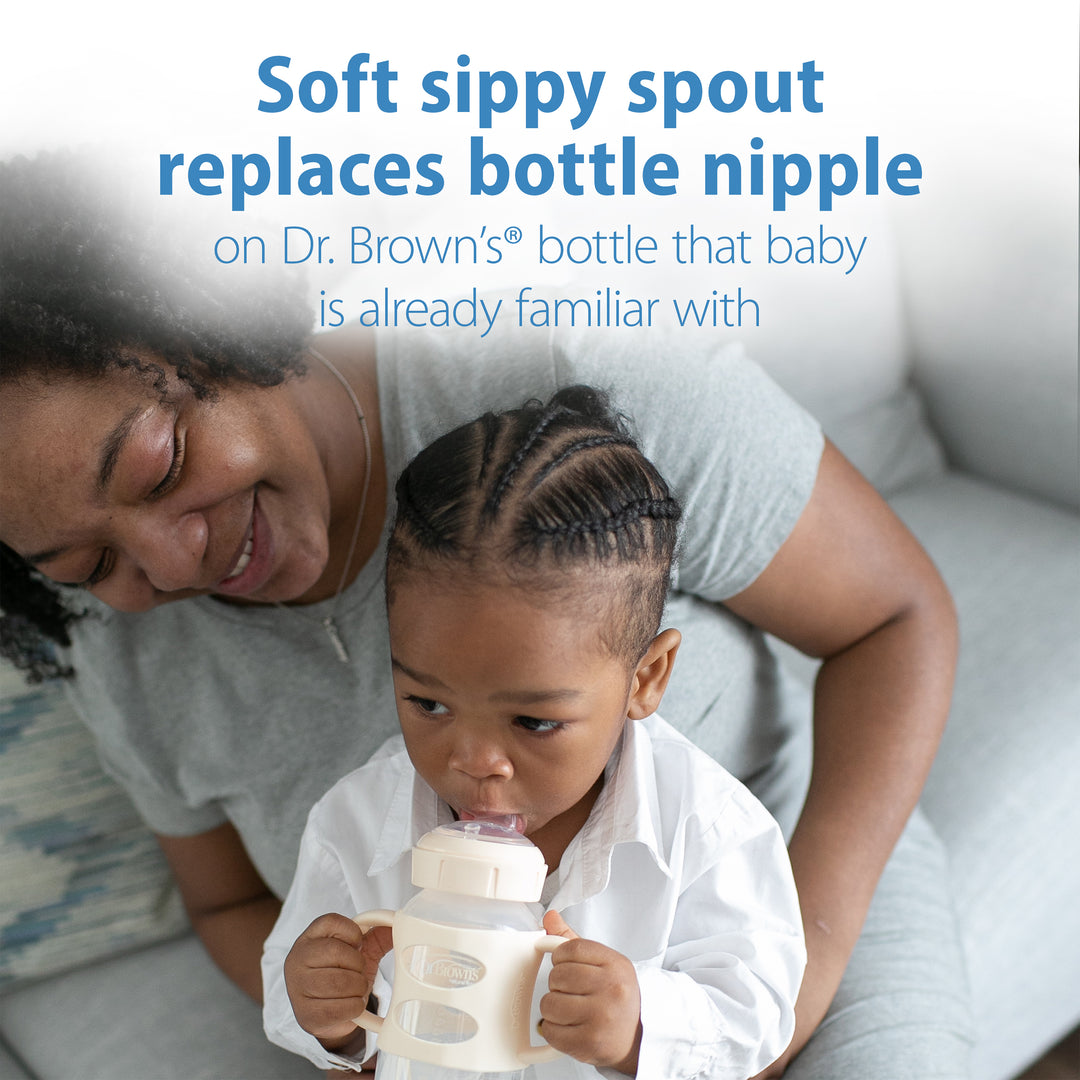 A toddler with braided hair is seated on a sofa, drinking from the Dr. Brown’s® Milestones™ Wide-Neck Sippy Spout Bottle with Silicone Handles, 9 oz/270 mL. Beside the child, an adult sits smiling and observing. The text on the image emphasizes how this Dr. Brown's bottle promotes drinking skills by seamlessly transitioning from a bottle nipple to its sippy spout design.