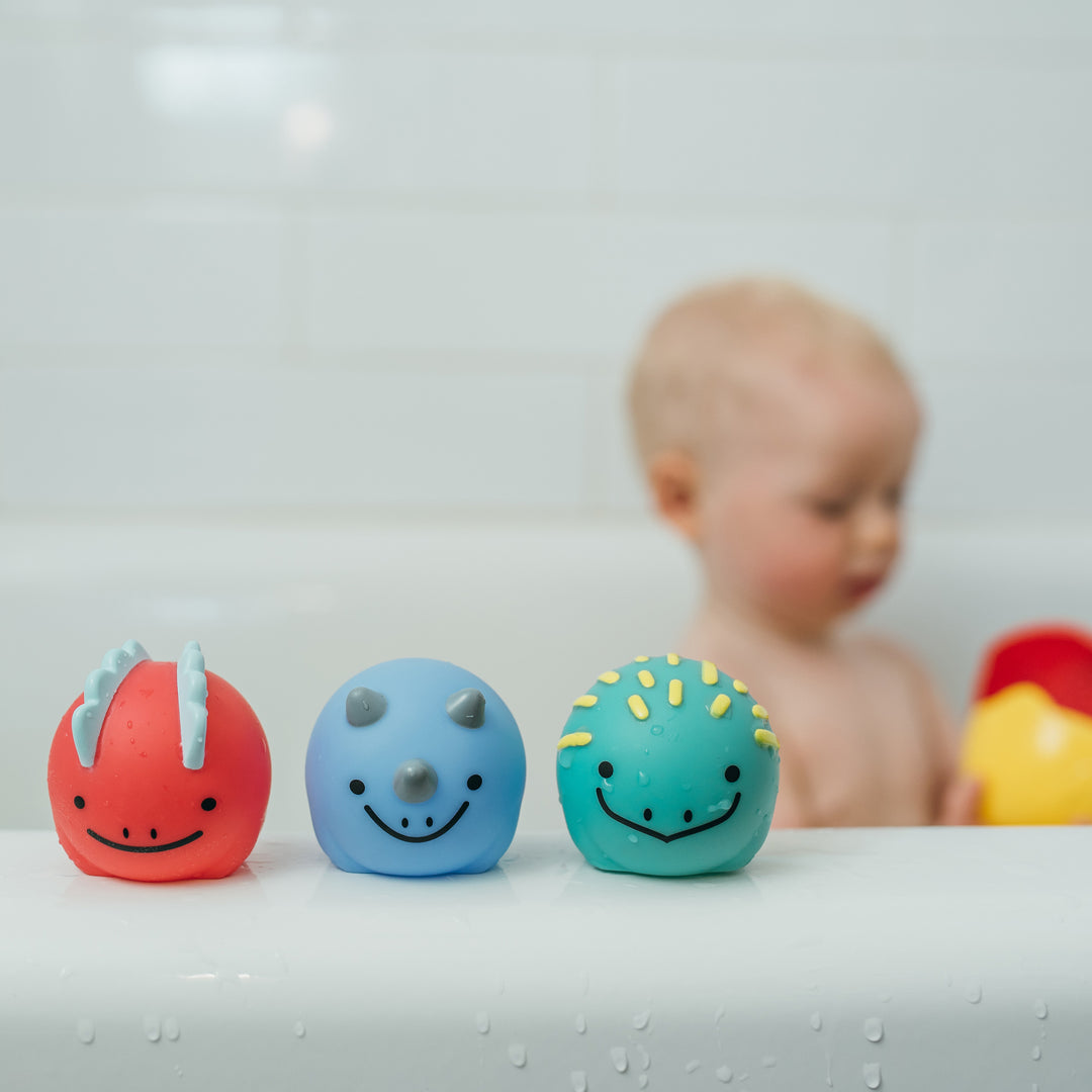 A child plays in a bathtub, surrounded by visual stimulation. In the foreground, three colorful dinosaur-themed baby bath toys from Dr. Brown's CleanUp™ Float & Hatch Dino Eggs set with smiling faces are lined up on the tub's edge: a red one with horns, a blue one with a nose horn, and a teal one with spikes—perfect nesting dino friends for endless fun.
