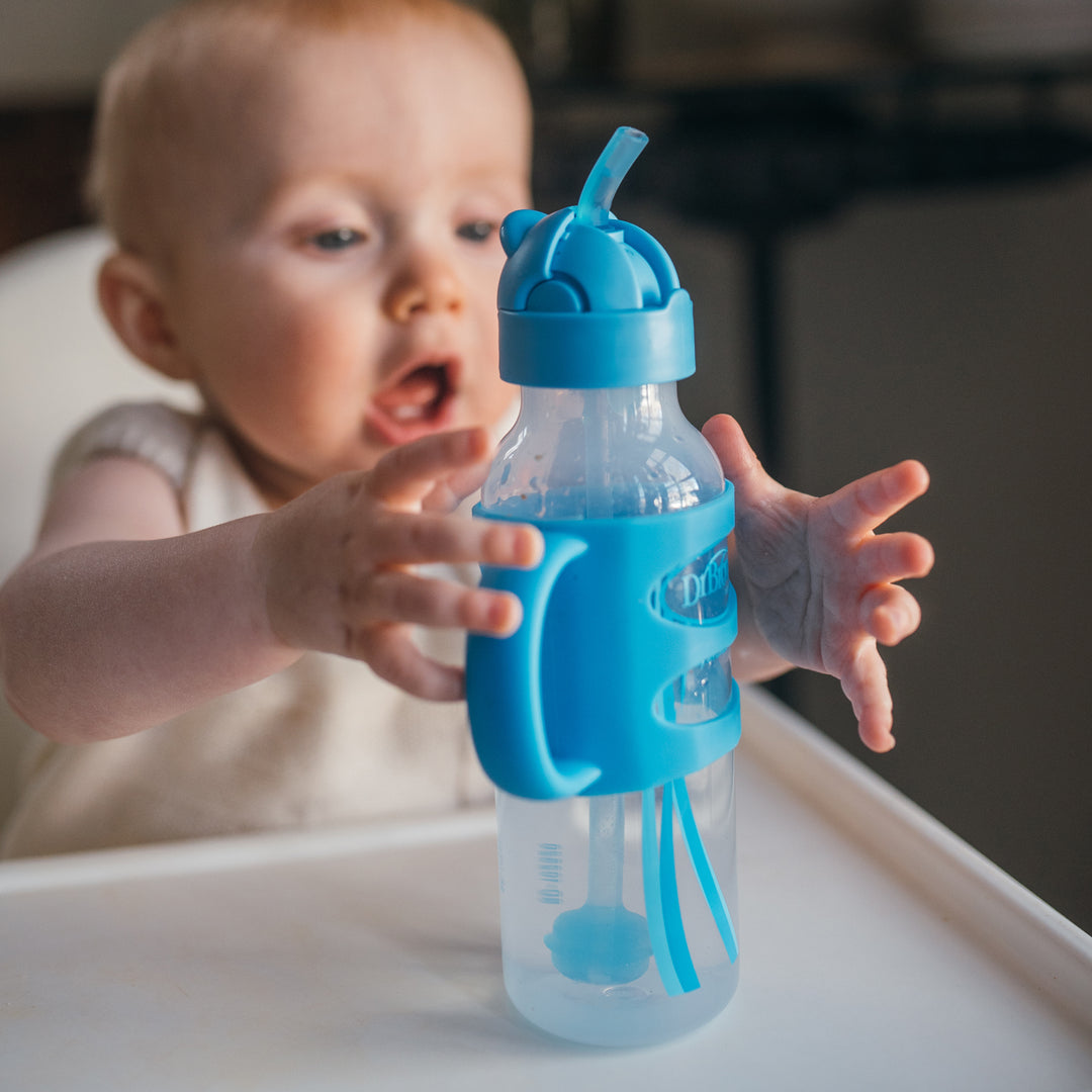 A baby eagerly reaches for the Dr. Brown’s™ Milestones™ Narrow Sippy Straw Bottle with Silicone Handles on a white high chair tray. This 8 oz/250 mL bottle features a flexible silicone weighted straw and side handles, making it perfect for supporting independent drinking. The baby's expression radiates excitement to grab the cup from Dr. Brown's collection.