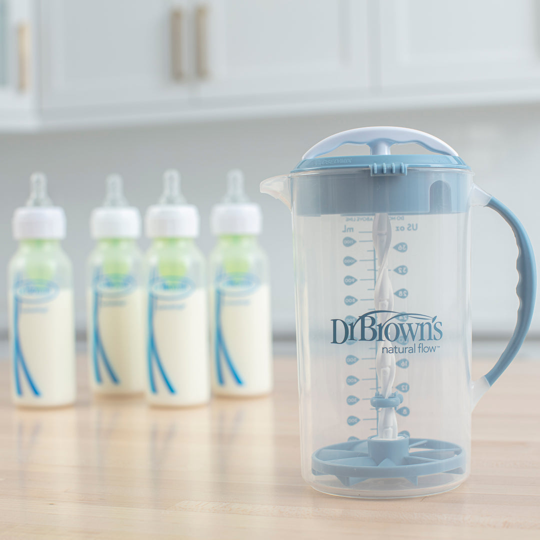 A Dr. Brown's Natural Flow® Formula Mixing Pitcher, featuring a clear plastic design with a blue handle and lid, rests on a wooden kitchen surface. In the background, four baby bottles filled with milk are blurred, showcasing the pitcher’s effectiveness in minimizing gas for more enjoyable feeding times.