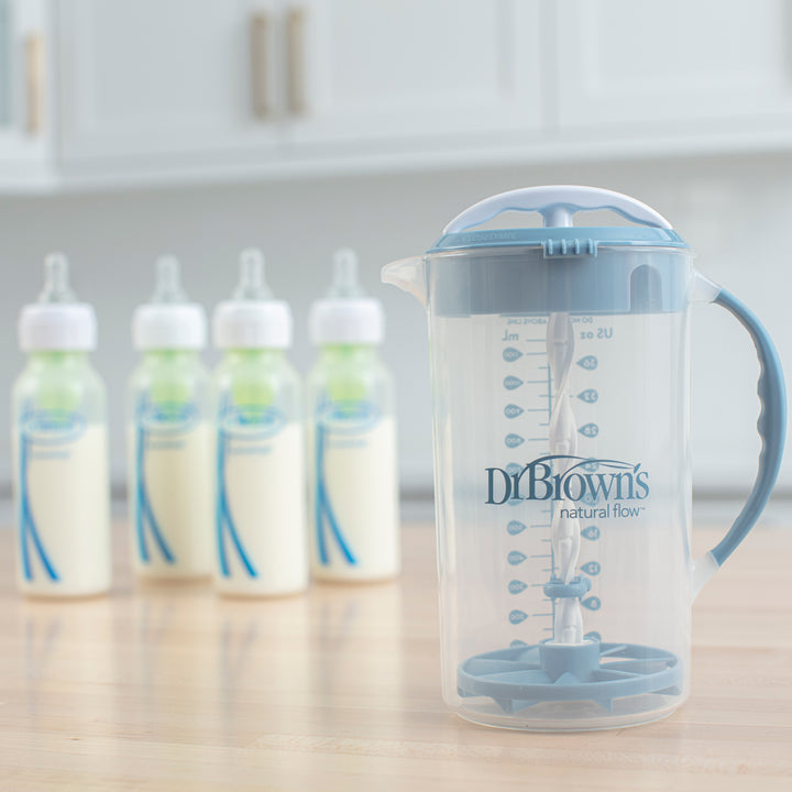 A Dr. Brown's Natural Flow® Formula Mixing Pitcher, featuring a clear plastic design with a blue handle and lid, rests on a wooden kitchen surface. In the background, four baby bottles filled with milk are blurred, showcasing the pitcher’s effectiveness in minimizing gas for more enjoyable feeding times.