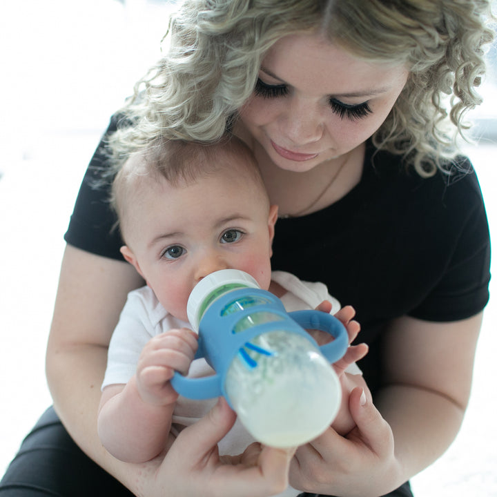 A woman with curly hair holds a baby in her lap, helping the child develop independent drinking skills. The baby grasps the Dr. Brown’s® Milestones™ Wide-Neck Silicone Handles bottle, and they both appear calm and focused, set against a softly blurred background.