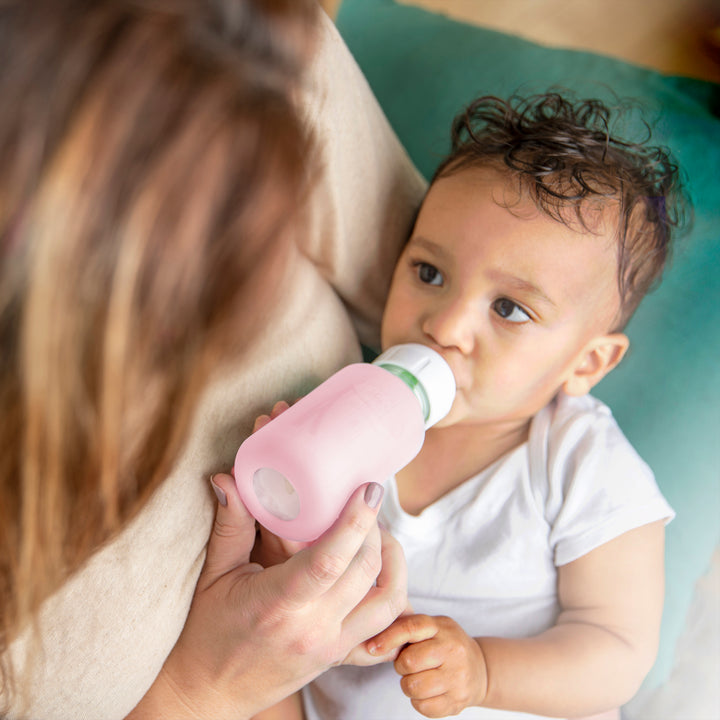 A baby in a white onesie rests comfortably, sipping from a pink Dr. Brown’s Natural Flow® Options+™ Narrow Glass Bottle Silicone Sleeves, held by an adult with long brown hair. With curly hair and tiny fingers gripping the bottle, the baby gazes up contentedly against a soft green backdrop.