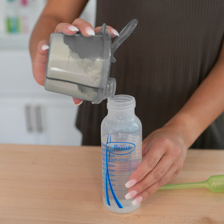 A person is preparing for baby feeding by pouring powdered formula from a grey container into a Dr. Brown's 9 oz bottle filled with water. The bottle rests on a light-colored wooden surface, with Dr. Brown’s® Baby Formula Dispenser and a green scoop nearby.