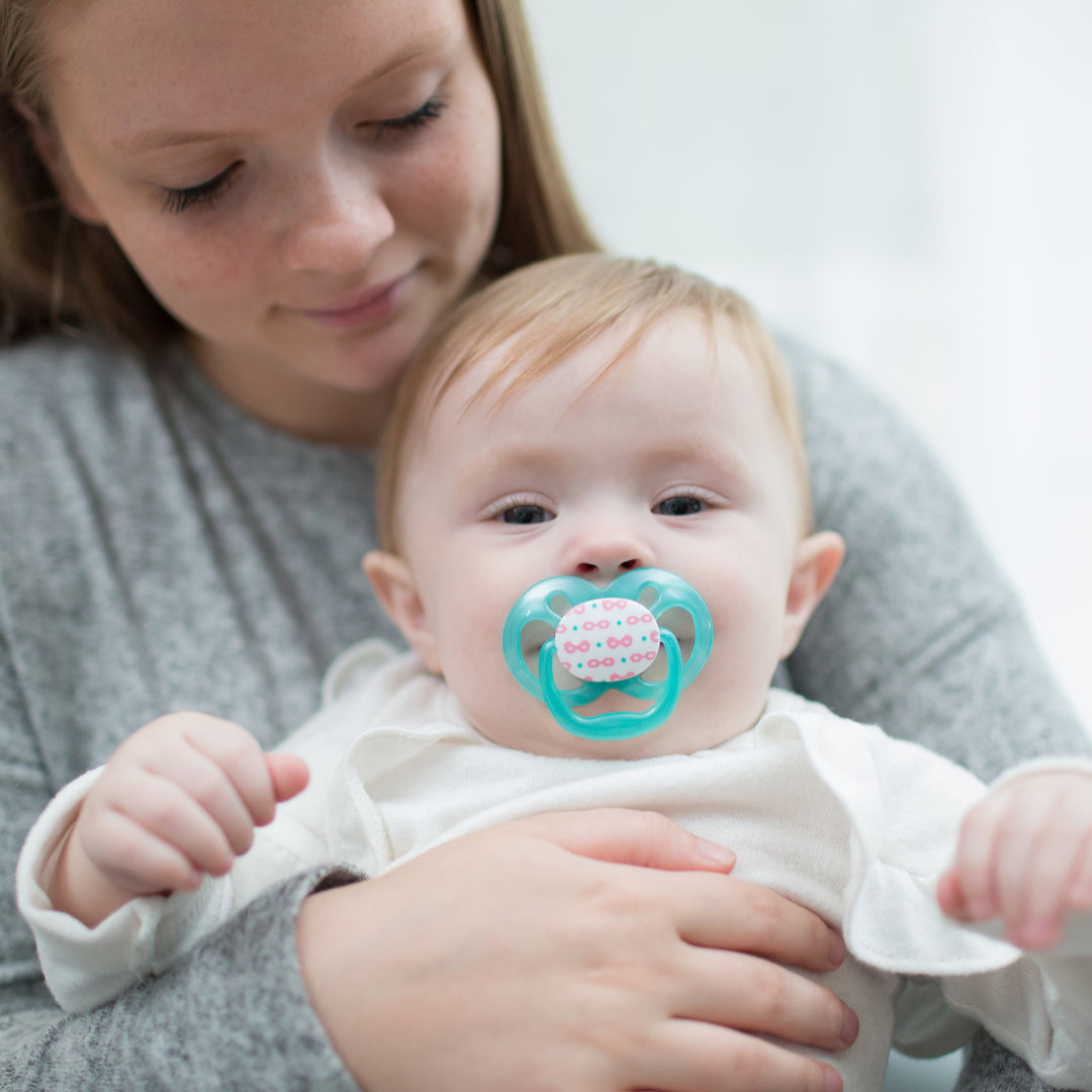 A woman gently cradles a baby with a Dr. Brown’s™ Advantage™ Pacifier. The baby, nestled in her arms, gazes at the camera. Both are dressed in soft, light-colored clothing, set against a blurred background that creates a calm, symmetrical setting.