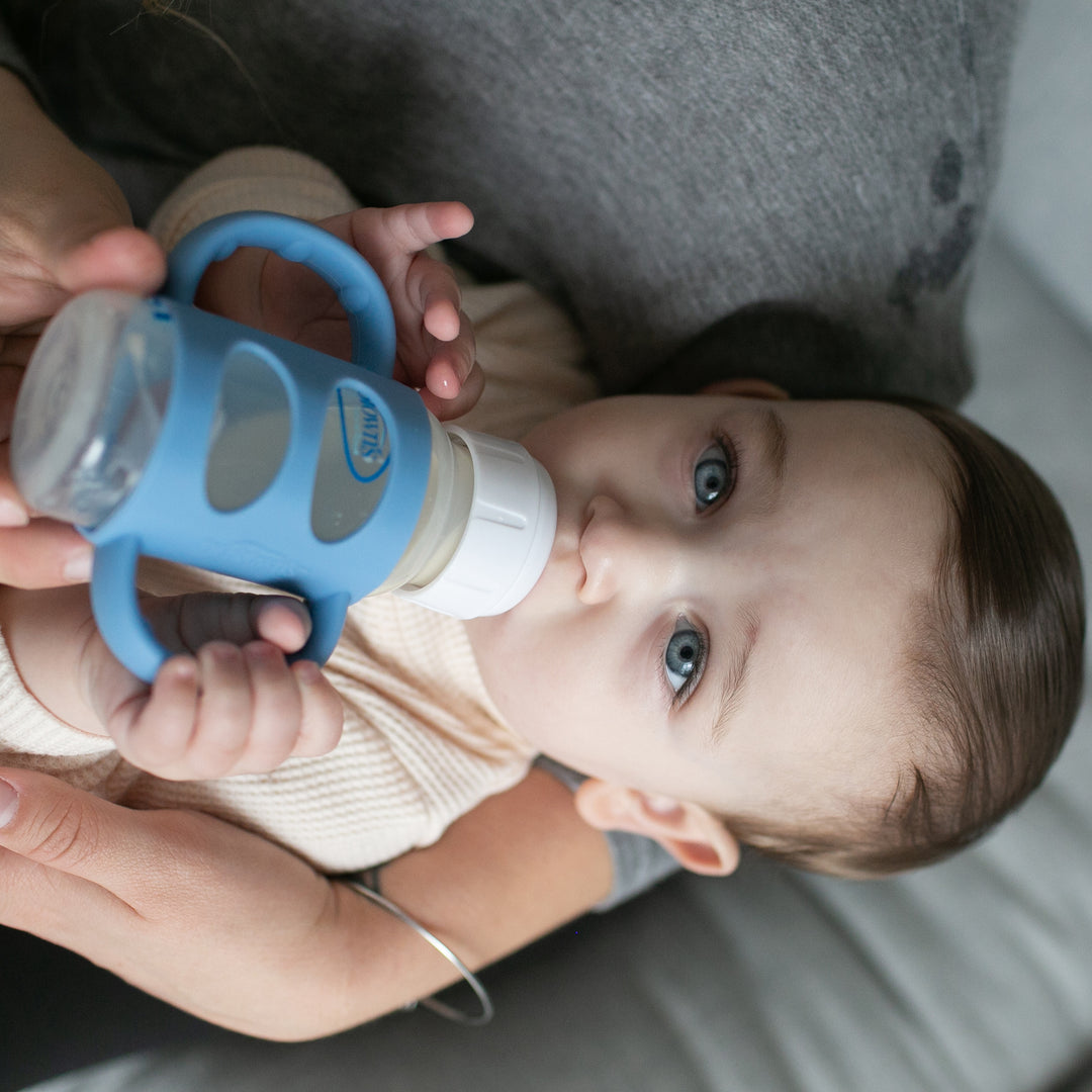 A baby with blue eyes is lying down, practicing independent drinking skills while sipping from a bottle equipped with Dr. Brown’s® Milestones™ Narrow Silicone Handles. The adult gently supports the child, who is dressed in a light-colored outfit, ensuring a comforting experience.

