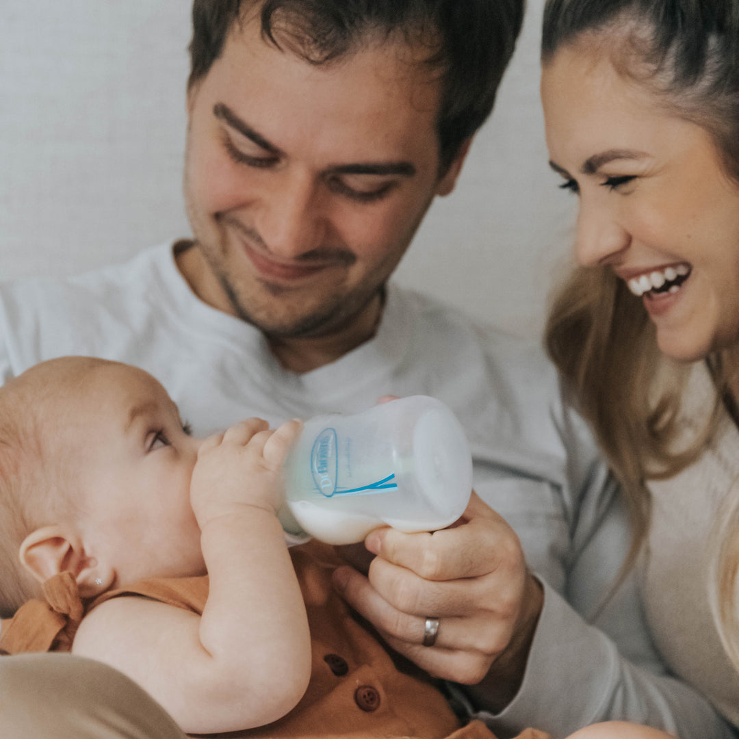 A happy couple sits together, smiling at their baby who is drinking from a Dr. Brown's Natural Flow® Anti-Colic Options+™ Wide-Neck Baby Bottle, featuring a Level 1 Slow Flow Nipple. The man holds the bottle while the woman leans in, with all three appearing joyful and affectionate.