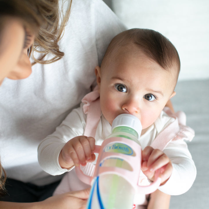 A baby with short brown hair is drinking from a Dr. Brown's Narrow Bottle held by an adult. The baby, dressed in a white shirt and light pink outfit, gazes up with large eyes. Dr. Brown’s® Milestones™ Narrow Silicone Handles would help nurture independent drinking skills while the adult's face remains partially visible, showing brown hair.