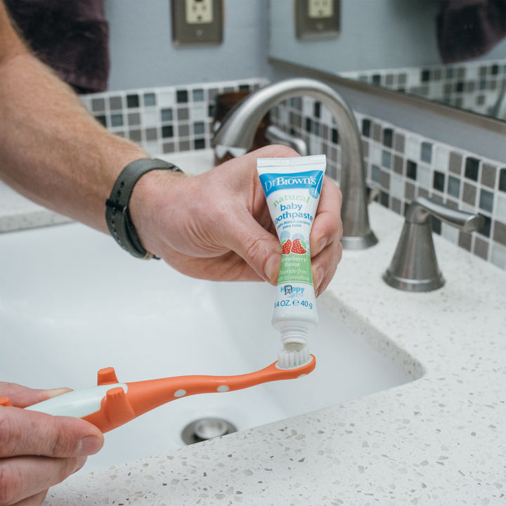 A person is applying Dr. Brown’s™ Fluoride-Free Baby Toothpaste, Strawberry, 1.4oz/40g, 1-Pack onto an orange and white toothbrush over a bathroom sink to promote dental hygiene. The background includes a tiled wall, faucet, and light switch.