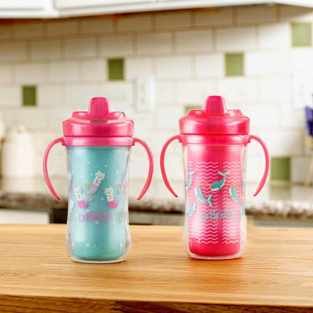 On a wooden kitchen counter, two vibrant children's sippy cups with handles are displayed. The left cup is part of the Dr. Brown’s Milestones™ Insulated Hard Spout Sippy Cup 2-Pack and features a blue design with unicorn motifs, while the right cup is adorned in pink with mermaid designs. Both cups come equipped with bite-resistant hard spouts and have pink lids set against a tiled backsplash.