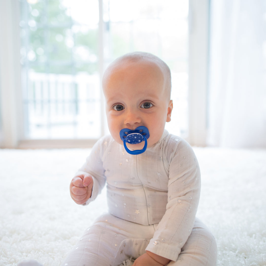 A baby with a blue Dr. Brown’s™ Advantage™ Pacifier from a 2-count pack is seated on a soft white carpet in a bright room. The baby wears a light-colored outfit and gazes towards the camera, while natural light filters in through sheer curtains on the window in the background.