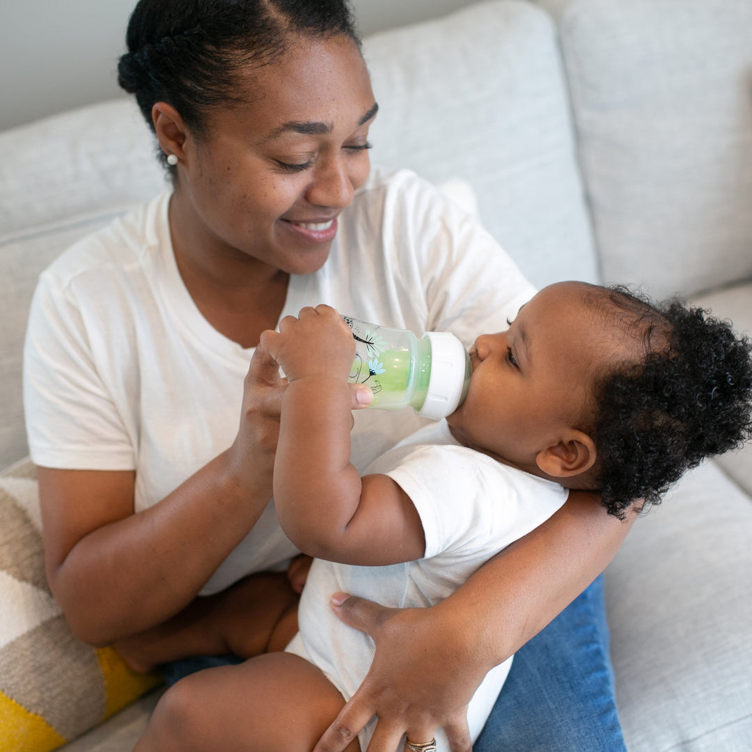 A woman is lovingly holding a baby, who is drinking from a Dr. Brown’s Natural Flow® Anti-Colic Options+™ Wide-Neck Baby Bottle. They are both sitting on a light-colored couch. The woman, smiling in a white t-shirt, holds the child securely in her lap as they enjoy this tender moment.