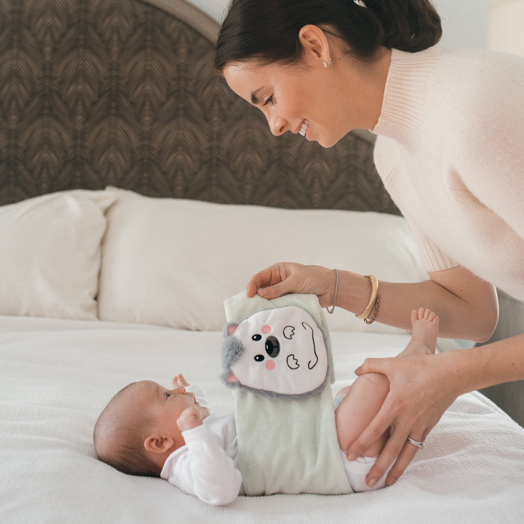 A woman smiling at a baby lying on a bed is using a cute animal-themed diaper alongside Dr. Brown’s™ Infant Gripebelt™ Colic Relief Belt by Dr. Brown's for natural gas and colic relief. Both appear happy and relaxed in a cozy bedroom setting.